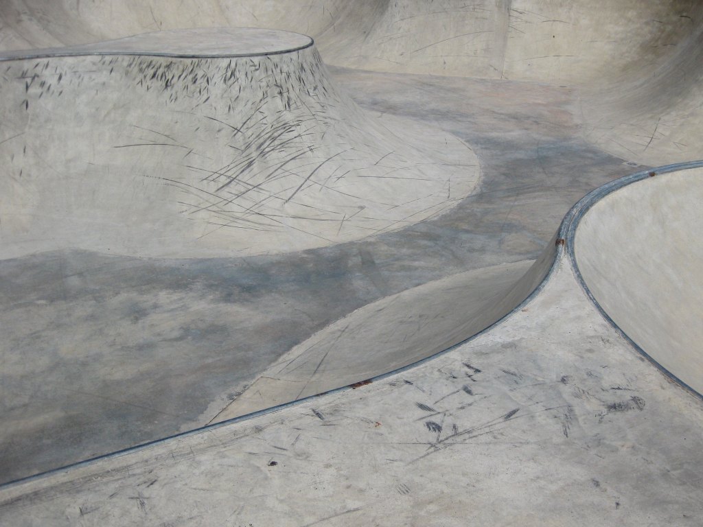 A newly built skatepark showing concrete bowls and transitions to metal rails, all looking a little like an abstract image of an alien wind swept landscape. Black tyre marks are beginning to build up showing where riders are most commonly starting to ride