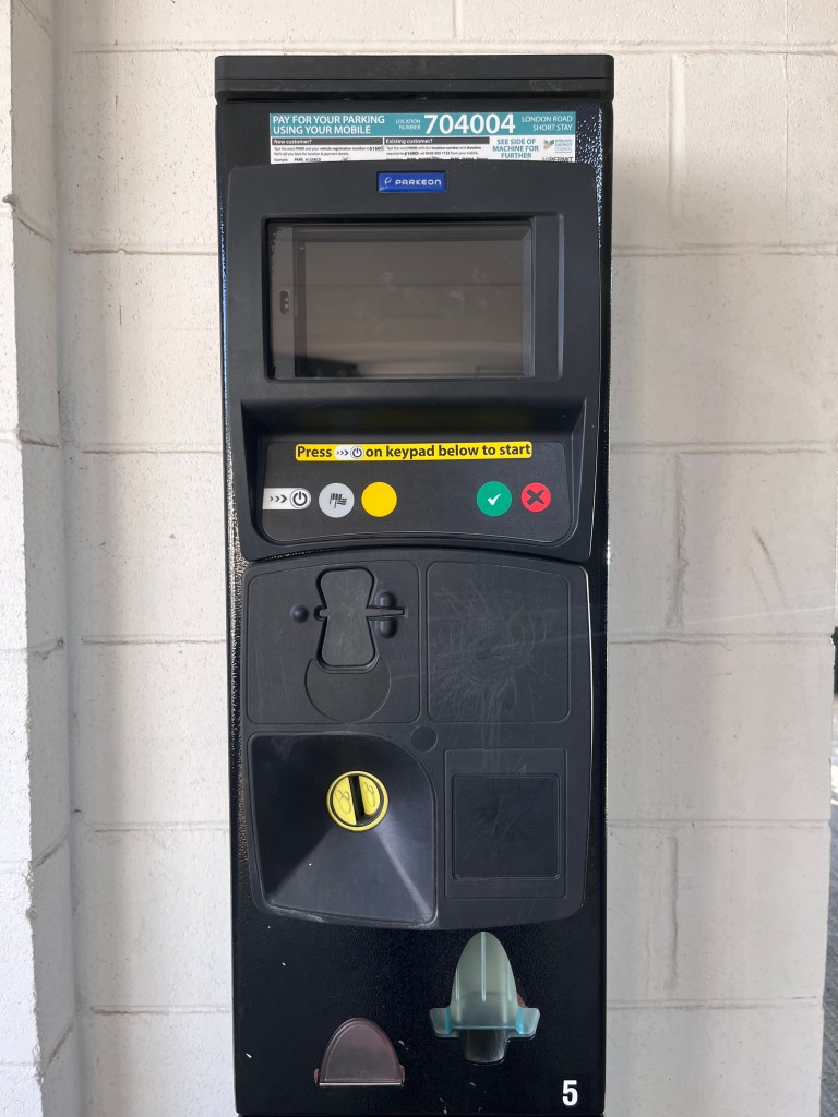 A photo of a black parking meter machine It has a dark blank digital screen, a green button with a tick, a red button with a cross, and random power, flag, and yellow buttons, over a coin slot. On the top is a sticker with instructions. 