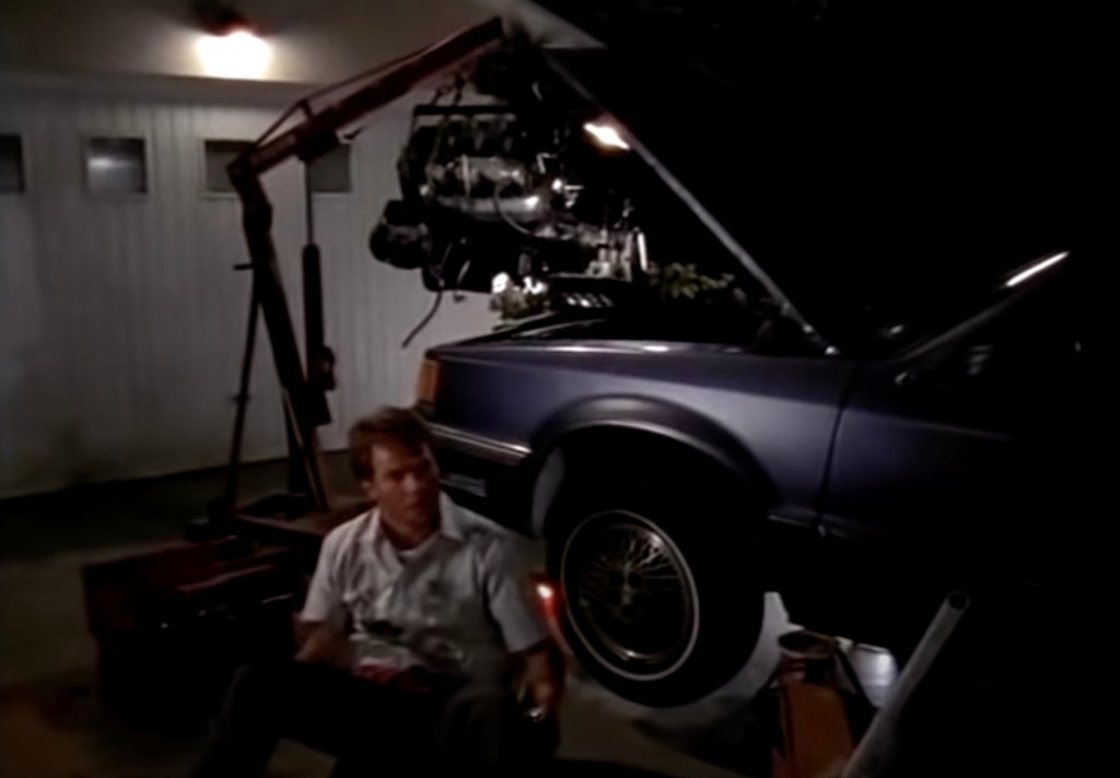A man in a white shirt sits on the floor after emerging from under a car that is jacked up with the engine removed. He is oily and angry looking after being interrupted from fixing the car