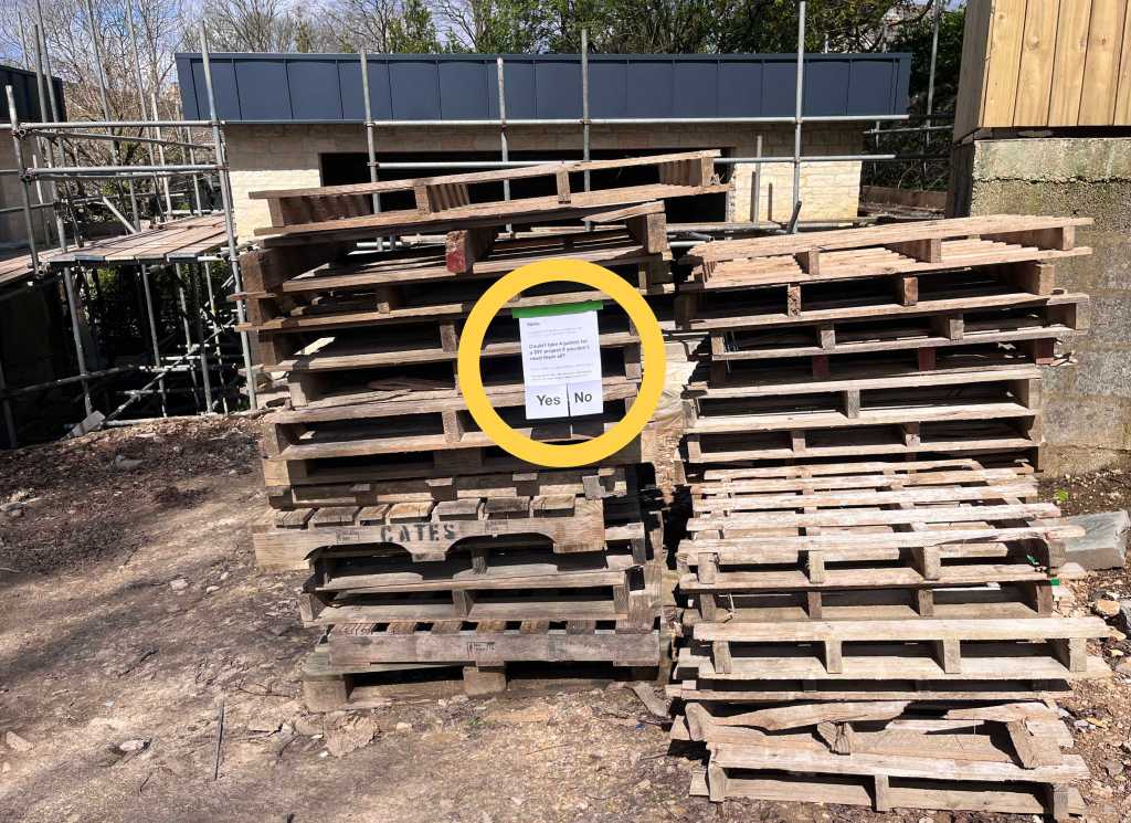 A wide photo of a building site showing a large pile of wooden pallets and an A4 page stuck to them. A yellow circle highlights the A4 page. A big friendly yes no option is clearly visible at the bottom of the page