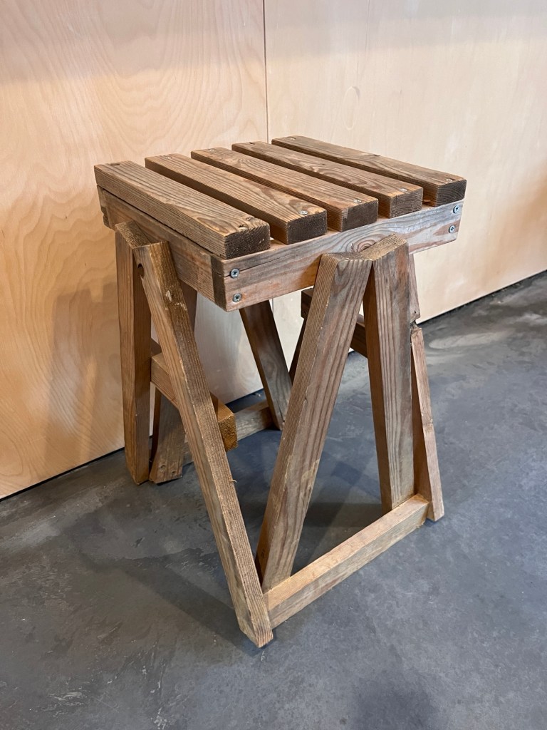 A simple rustic looking angular stool made of off cuts of wood batten. The legs are capital A shaped and the seat is slatted. It’s very basic yet oddly architectural and practical looking. 