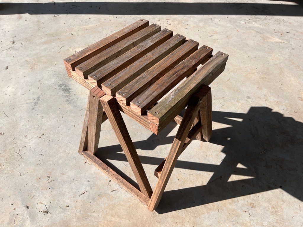 A high contrast photo of my simple rustic looking angular stool made of off cuts of wood batten. The legs are capital A shaped and the seat is slatted. It’s very basic yet oddly architectural and practical looking. 